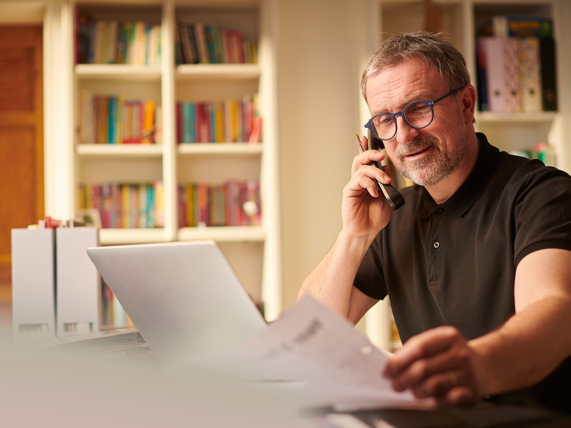 Older man sitting at a desk while talking on the phone and reviewing his laptop and paperwork.